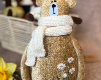 May include: A plush brown bear wearing a white knitted scarf and embroidered with white flowers. The bear is sitting on a light brown surface.