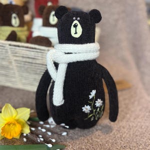 May include: A brown knitted teddy bear wearing a white scarf and embroidered white flowers. The bear has black eyes and a black nose.