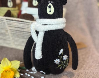 May include: A brown knitted teddy bear with a white scarf and embroidered white flowers. The bear has black eyes and a black nose.