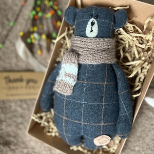 May include: A plaid blue teddy bear with a brown knitted scarf. The bear is sitting in a brown box with shredded paper.