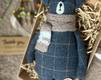 May include: A plaid blue teddy bear with a brown knitted scarf. The bear is sitting in a brown box with shredded paper.