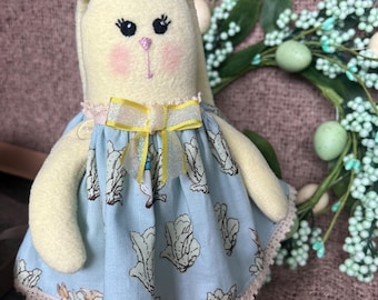 Könnte beinhalten: Ein gelber Plüschhase, der ein blaues Kleid mit weißer Spitzenborte und einer gelben Schleife trägt. Das Kleid hat ein Muster aus grünen Kohlblättern. Der Hase sitzt auf einer rosa gestrickten Oberfläche.