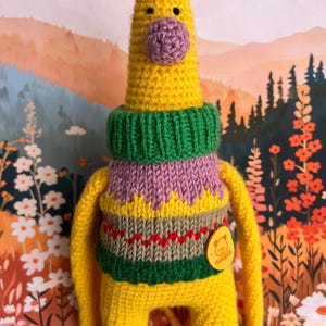 May include: A handmade yellow crocheted giraffe doll wearing a green and purple sweater. The giraffe has small brown horns and a purple nose. A small yellow tag says "Handmade". The background features a colorful floral design.