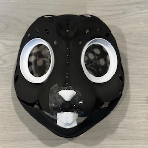 May include: Black furry mask with white eye and nose details. The mask has a textured surface and is designed to resemble an animal.