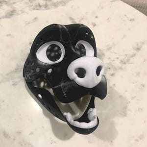 May include: A black and white 3D-printed mask of a canine or similar animal. The mask features large, open eye sockets, a white snout, and a wide-open mouth with white teeth. The mask has a textured surface and is designed to be worn.