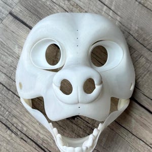 Könnte beinhalten: Weiße, unfertige Felltiermaske mit offenem Mund und sichtbaren Zähnen. Die Maske hat zwei große Augenlöcher und eine kleine Nase.