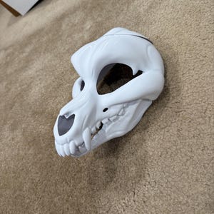 May include: A white, stylized skull mask with large eye sockets and prominent teeth. The mask has a smooth, glossy finish and is designed to be worn. The eye sockets and nose area are dark gray. The mask is likely for costume or cosplay purposes.