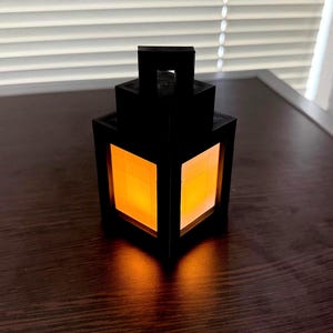 May include: A black lantern with a square design and a warm, orange glow emanating from within. The lantern has a rectangular handle at the top and is sitting on a dark wooden surface. The light creates a soft shadow.