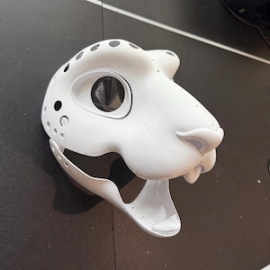 May include: White furry animal mask with a large open mouth and a black eye hole. The mask is made of a smooth, hard material and has a textured surface.