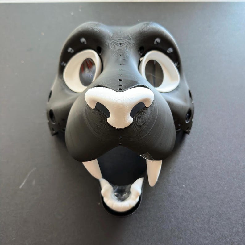 May include: A black and white 3D printed animal mask with a large open mouth and sharp teeth. The mask has white eyes, a white nose, and white teeth.