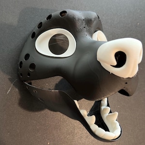 Puede incluir: Una máscara de animal de piel negra y blanca con la boca abierta y dientes visibles. La máscara tiene una nariz blanca y ojos grandes blancos.