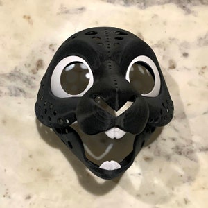 May include: Black and white furry animal mask with a detailed face and large eyes. The mask is made of a textured material and has a hole for the mouth.