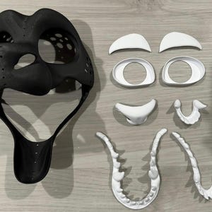 May include: A black 3D printed mask with white 3D printed facial features, including eyes, eyebrows, a nose, and teeth. The mask is designed for use in cosplay or costume making.