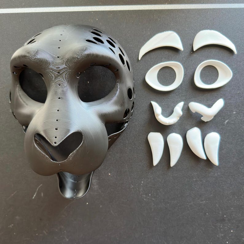 May include: A black 3D printed animal mask with white plastic pieces for the eyes, nose, and eyebrows. The mask is designed to look like a leopard or cheetah.