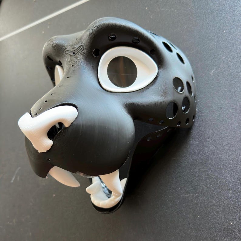 May include: A black and white furry animal head mask with a large white eye and a white nose. The mask is made of a textured material and has a mouth with white teeth.
