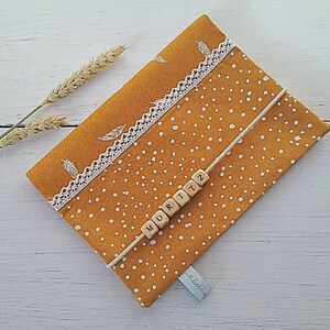 Op de afbeelding: Een gele stoffen tas met witte stippen en witte kantversiering. De tas heeft een wit koord met houten letterparels die "MORITZ" op de voorkant spellen.