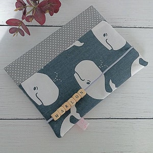 Op de afbeelding: Een stoffen boekomslag met een walvismotief in blauw- en wittinten. De omslag is vastgezet met een grijze elastische band en heeft de naam "MARLON" gespeld met houten letterkralen. Een grijze stof met stippen is zichtbaar.