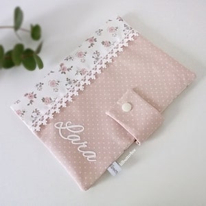 Op de afbeelding: Een roze stoffen babyboek met witte stippen en een bloemenpatroon. Het boek heeft een witte kantversiering en de naam "Lara" is geborduurd op de voorkant. Het boek heeft een klein zakje aan de zijkant met een witte knoop.