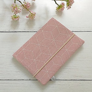 Op de afbeelding: Een rechthoekige, roze stoffen hoes met een wit geometrisch kubuspatroon. Een beige elastiek sluit de hoes. De hoes is waarschijnlijk voor een tablet of e-reader. Een takje roze bloemen ligt op de achtergrond.