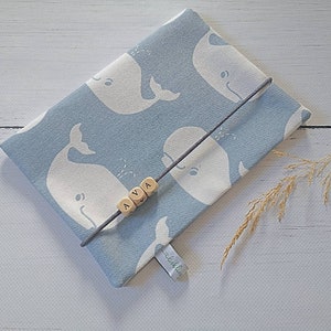 Op de afbeelding: Een lichtblauwe stoffen tas met een wit walvispatroon. De tas heeft een grijs trekkoord en houten kralen met de letters "AVA" erop.