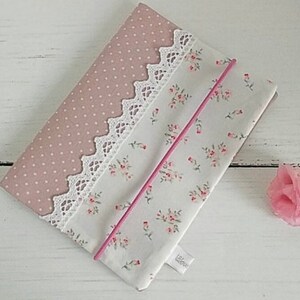 Op de afbeelding: Een roze en witte stoffen tas met een bloemenpatroon en kantversiering. De tas heeft een roze rits en een wit label met het woord "Little" erop.