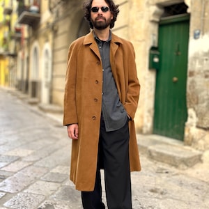 May include: A long, tan overcoat with a button-up front. The coat is worn over a dark gray button-down shirt and black pants. The person is wearing black shoes and sunglasses. The background is a cobblestone street and buildings.