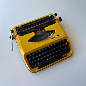 Può includere: Una macchina da scrivere Brother 440TR giallo brillante con tasti neri e un tasto tab rosso. La macchina da scrivere ha un design classico con un carrello in metallo e una disposizione della tastiera. Il logo Brother è visibile sulla parte anteriore.