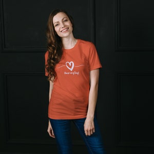 May include: A woman wearing a coral colored t-shirt with a white heart and the text "Have a nice day!" printed on the front. She is standing in front of a black wall.