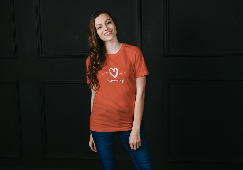 May include: A woman wearing a coral colored t-shirt with a white heart and the text "Have a nice day!" printed on the front. She is standing in front of a black wall.