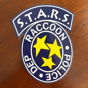 May include: A blue and white sticker featuring the text "S.T.A.R.S. RACCOON DEP POLICE" in a circular design. The center of the circle has three yellow stars.