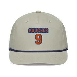 May include: A light gray baseball cap with the name "BOUCHER" embroidered in blue and orange, with the number "9" in orange. The cap has a navy blue rope detail along the brim.
