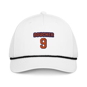 May include: White baseball cap with the name "BOUCHER" embroidered in orange and blue, and the number "9" in orange. The cap has a black rope detail along the brim.
