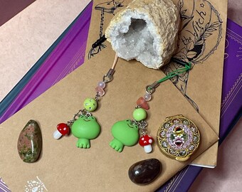 May include: Two green frog charm keychains with red mushroom accents and crystal beads. The keychains are displayed on a brown surface with a crystal and a small decorative box.