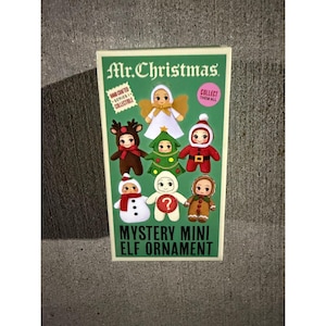 May include: A rectangular Mr. Christmas Mystery Mini Elf Ornament box featuring a green background with illustrations of six small, stylized holiday-themed figures including an angel, reindeer, Christmas tree, Santa, snowman, and gingerbread person.