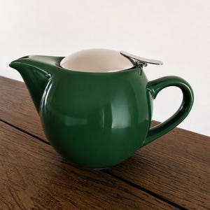 May include: A dark green ceramic teapot with a rounded body, a curved handle, and a pointed spout. The teapot features a polished silver-colored metal lid with a small, flat lever attached to the side for opening and closing the top securely.