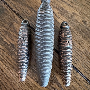 May include: Three decorative pine cone shaped objects. The largest is gray, and the two smaller ones are bronze with dark accents. All three have a loop at the top for hanging. The objects are on a wooden surface.