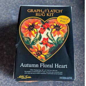 May include: A Graph N' Latch rug kit box titled Autumn Floral Heart. The black box features a heart-shaped graphic of orange, yellow, and red flowers with green leaves. Text indicates a finished size of 30 inches by 27 inches.