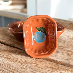 Puede incluir: Dos pequeños cuencos de cerámica de color terracota. Un cuenco está enfocado, con un diseño rectangular en relieve con una fruta azul y un detalle de hoja verde. El otro cuenco está en el fondo. Los cuencos están sobre una superficie de madera.