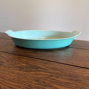 May include: A shallow, oval-shaped baking dish with a vibrant turquoise exterior and a smooth, cream-colored interior. The dish features two integrated handles on opposite sides, providing a simple and functional design for oven-to-table use.