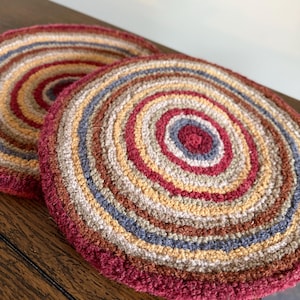 May include: Two round, crocheted coasters featuring concentric circles in a palette of deep burgundy, muted blue, warm tan, and cream. The textured yarn creates a thick, circular pattern that radiates from the center to the outer edge.