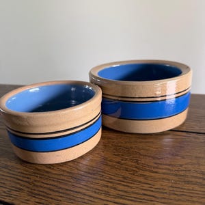 May include: Two ceramic bowls with a tan exterior, a blue interior, and a blue stripe around the middle. One bowl is slightly smaller than the other. The bowls are sitting on a wooden surface.