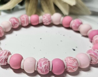 May include: A stretch bracelet composed of round beads in various shades of pink, including solid light pink, solid dark pink, and marbled pink and white patterns, separated by small, thin, flat white disc spacers arranged in a repeating sequence.