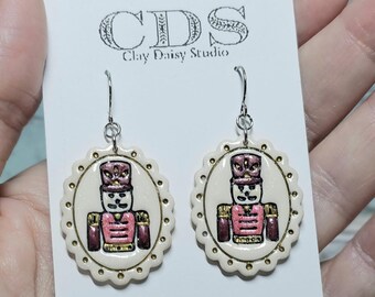 May include: A pair of oval-shaped earrings featuring a painted nutcracker design in pink, gold, and dark red on a cream background with a scalloped edge. The earrings have surgical stainless steel hooks and measure 5.08 cm in length.