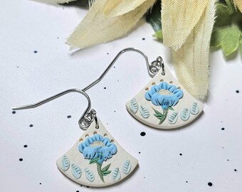 May include: A pair of fan-shaped dangle earrings featuring a cream-coloured base with a raised light blue flower design, green stems, and small blue leaf accents. The earrings are attached to silver-toned metal hooks for pierced ears.