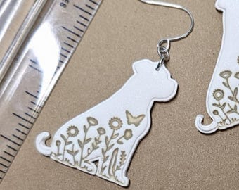 May include: A pair of white drop earrings shaped like sitting dogs with etched floral and butterfly designs. The earrings feature silver-toned metal hooks and are positioned next to a clear plastic ruler showing a measurement of 5 cm.