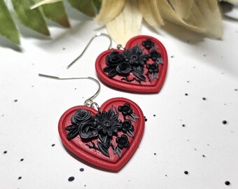 May include: A pair of heart-shaped drop earrings featuring a vibrant red base with intricate, raised black floral embellishments. Each earring is attached to a silver-toned metal hook, showcasing a detailed sculpted design of roses and leaves.