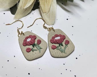 May include: A pair of drop earrings featuring a beige, textured, trapezoidal base with a raised floral design. The flowers are dark pink with white centres and green stems. The earrings are attached to gold-toned metal hooks for pierced ears.