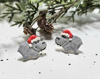 May include: A pair of stud earrings shaped like grey hippopotamuses, each wearing a red Santa hat with a white textured trim. The hippos have a smooth grey finish, visible white teeth, and are positioned side by side on a white surface.
