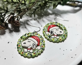 May include: A pair of dangle earrings featuring a circular green wreath design with a white dog character wearing a red Santa hat. The earrings have silver-toned metal hooks and a glossy finish, depicting the character centred within the wreath.
