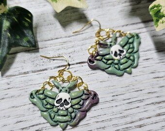 May include: A pair of dangle earrings featuring moth-shaped charms with green and purple iridescent wings and a white skull detail in the centre. The charms are attached to gold-toned metal hooks by small circular gold-toned metal jump rings.